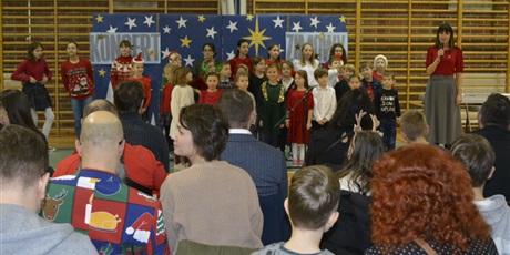 Powiększ grafikę: Grupka kilkunastu dzieci z chóru szkolnego wraz z nauczycielką muzyki stoją na scenie sali gimnastycznej.