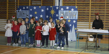 Powiększ grafikę: Grupka kilkunastu dzieci ze świetlicy szkolnej wraz z nauczycielką grającą na syntezatorze śpiewają kolędę na sali gimnastycznej.