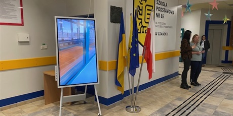 Powiększ grafikę: Korytarz szkolny: wejście główne. Na korytarzu stoi duży telewizor do projekcji, flagi Polski, Unii Europejskiej i Ukrainy.