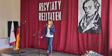 Powiększ grafikę: Antonina Oleszko-uczennica stoi na środku sceny przed mikrofonem i recytuje z ekspresyjną gestykulacją wiersz