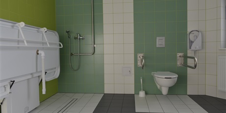 Powiększ grafikę: Łazienka dla osób niepełnosprawnych: na ścianach i podłodze kafle w kolorach białym, zielonym i szarym. Z lewej strony umieszczone jest łóżko do kąpieli i bateria prysznicowa. Z prawej strony wisi wc.