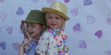 Powiększ grafikę: Dwie uśmiechnięte dziewczynki w kapeluszach i kolorowych łańcuchach na szyi pozują do zdjęcia odwrócone do siebie plecami