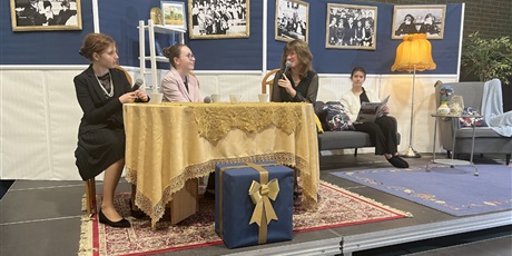 Powiększ grafikę: Na scenie w sali gimnastycznej uczennice grają swoje role w przedstawieniu. 3 uczennice siedzą przy stole, jedna siedzi na kanapie.