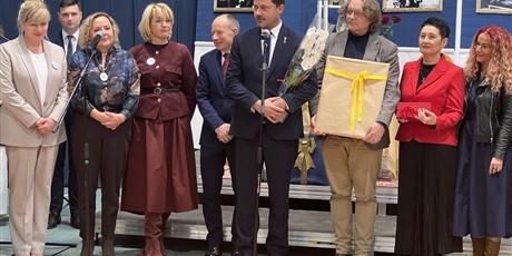 Powiększ grafikę: kobiety i mężczyźni, dyrektorzy szkół stoją na scenie, przed nimi 2 mikrofony. jeden mężczyzna trzyma kwiaty, a drugi prezent 