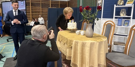Powiększ grafikę: Pani dyrektor zdmuchuje świeczkę na torcie, który stoi na scenie na stole z żółtym obrusem. Z lewej stronie klęczy fotograf i za nim stoi mężczyzna