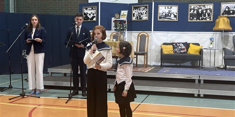 Powiększ grafikę: 2 dziewczynki stoją na scenie, jedna z mikrofonem, za nimi scena i  prowadzący uroczystość kobieta i mężczyzna 