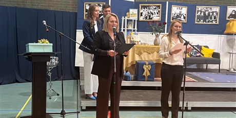 Powiększ grafikę: Dwie kobiety stoją przy mikrofonach na sali gimnastycznej, za nimi scena i dwoje młodych ludzi prowadzących uroczystość