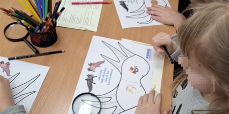 Powiększ grafikę: stół przy którym siedzą dzieci i rysują na kartkach zwierzątka, na stole leżą lupy i kredki oraz pojemnik z kredkami