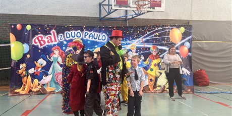 Powiększ grafikę: dzieci na sali gimnastycznej w strojach karnawałowych stoją pod plakatem, po środku stoi z mikrofonem klaun w czerwonej czapce