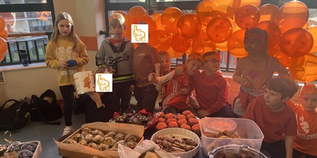 Powiększ grafikę: 9 dzieci z klas młodszych pozuje do zdjęcia. Za niemi pomarańczowe balony, przed nimi stół z ciastami