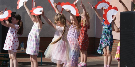 Powiększ grafikę: dzieci tańczą na scenie, trzymają biało-czerwone duże kwiaty z papieru