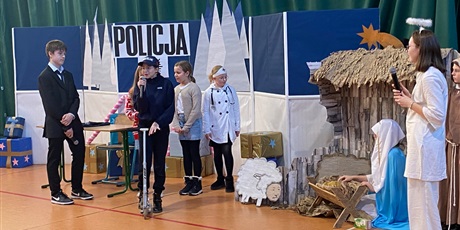 Powiększ grafikę: Na scenie policjantka na hulajnodze, lekarka w białym fartuchu, Maryja i Józef, w tle stajenka, dalej banner Policja oraz inni aktorzy i anioł z białą aureolą