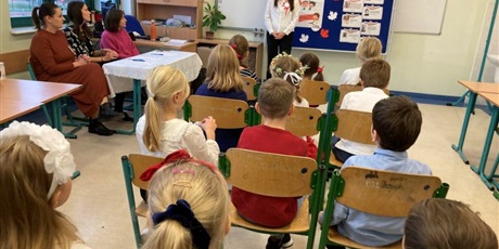 Powiększ grafikę: Dzieci siedzące tyłem na krzesełkach słuchają stojącego przed nimi nauczyciela. Z lewej strony siedzą trzy panie z komisji konkursowej
