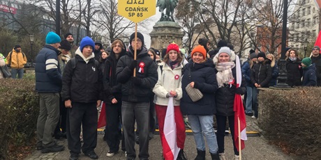 Powiększ grafikę: Młodzież i dorośli z flagą narodową i logiem naszej szkoły przed pomnikiem Piłsudskiego