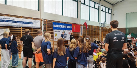 Powiększ grafikę: Dzieci i dorośli stoją tyłem na sali gimnastycznej i słuchają jak przewodniczący ogłasza wyniki