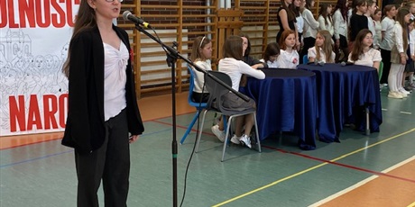 Powiększ grafikę: dziewczynka w okularach stoi przed mikrofonem na sali gimnastycznej. Z prawej strony przy 2 okrągłych stołach z granatowym obrusem siedzi 7 dziewczynek, a za nimi stoją dziewczęta z chóru.