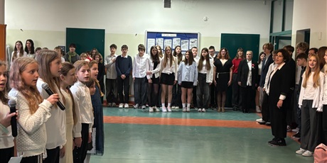 Powiększ grafikę: Uczniowie i nauczyciele stoją na holu szkoły przed salami i śpiewają hymn Polski