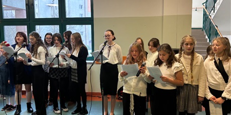 Powiększ grafikę: uczennice, dziewczynki z chóru szkolnego stoją przy mikrofonach z nutami na holu szkoły i śpiewają 