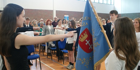 Powiększ grafikę: Uczniowie trzymają uniesione palce do ślubowania, stoją naprzeciwko sztandaru. Na sali uczniowie klas ósmych powtarzają słowa przysięgi.