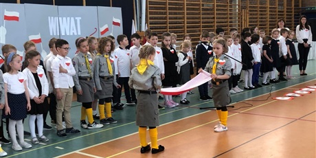 Powiększ grafikę: Dzieci stojące na scenie sali gimnastycznej. Dwie dziewczynki zwijają flagę Polski