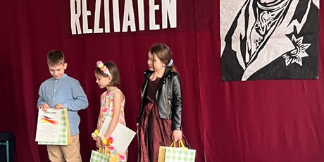 Powiększ grafikę: chłopiec i dwie dziewczynki stoją na scenie z dyplomami i torbami z nagrodami.