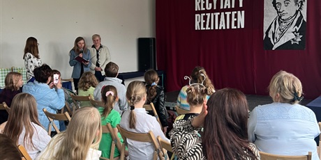 Powiększ grafikę: Dzieci z nauczycielami siedzą na krzesłach a 3 panie przemawiają do publiczności