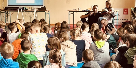 Powiększ grafikę: Dzieci siedzą na podłodze i słuchają mówiącego przez mikrofon gościa - pana siatkarza. Obok siatkarza siedzi dziewczynka z piłką.