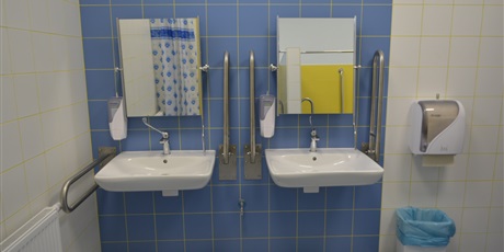 Powiększ grafikę: Dwie umieszczone obok siebie umywalka z lustrem: z lewej i prawej strony uchwyty dla osób niepełnosprawnych. Po prawej stronie na okaflowanej ścianie wisi pojemnik na papier do rąk.